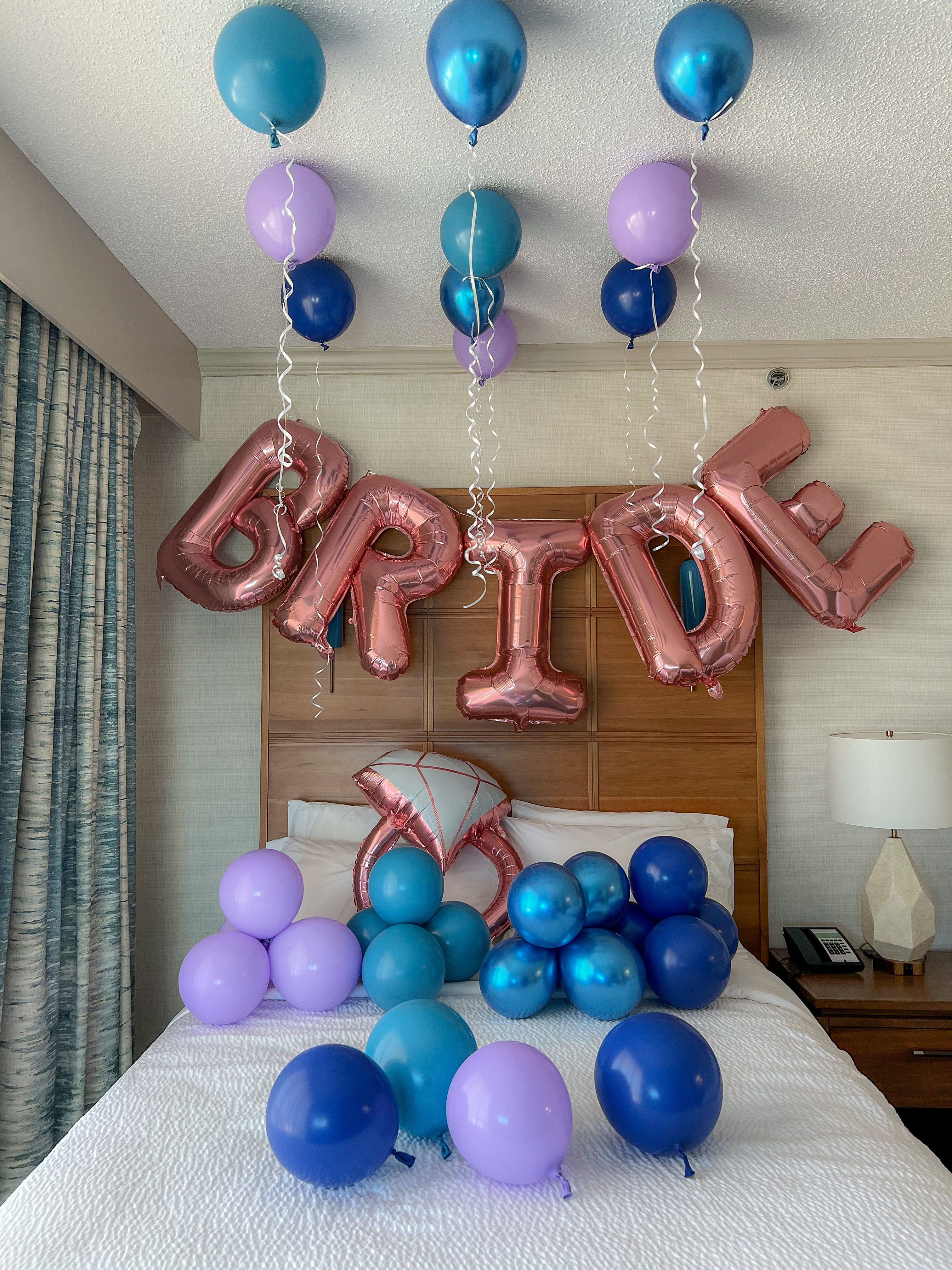 Hotel bedroom decorated for a bridal celebration with rose-gold foil 'BRIDE' letter balloons and teal, blue and purple latex balloons floating and clustered over the bed.
