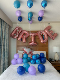Hotel bedroom decorated for a bridal celebration with rose-gold foil 'BRIDE' letter balloons and teal, blue and purple latex balloons floating and clustered over the bed.