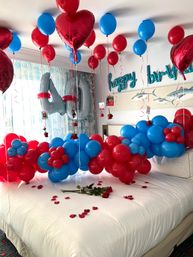 Festive bedroom scene with red and blue balloon clusters, heart-shaped red foil balloons, large silver '40' number balloons, teal 'happy birthday' foil letters on the wall and red rose petals scattered on the bed.