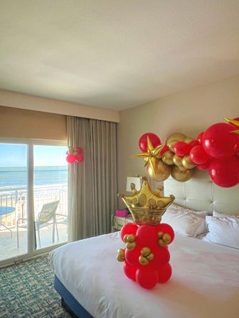Festive ocean-view hotel room with balcony and sliding glass door; bed topped by a red-and-gold balloon crown centerpiece and matching balloon garland over the headboard.