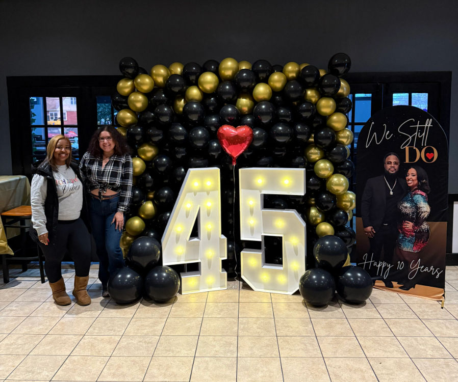 Indoor celebration backdrop with illuminated 45 marquee numbers, black-and-gold balloon wall, a red heart balloon and two smiling guests posing beside an anniversary sign.