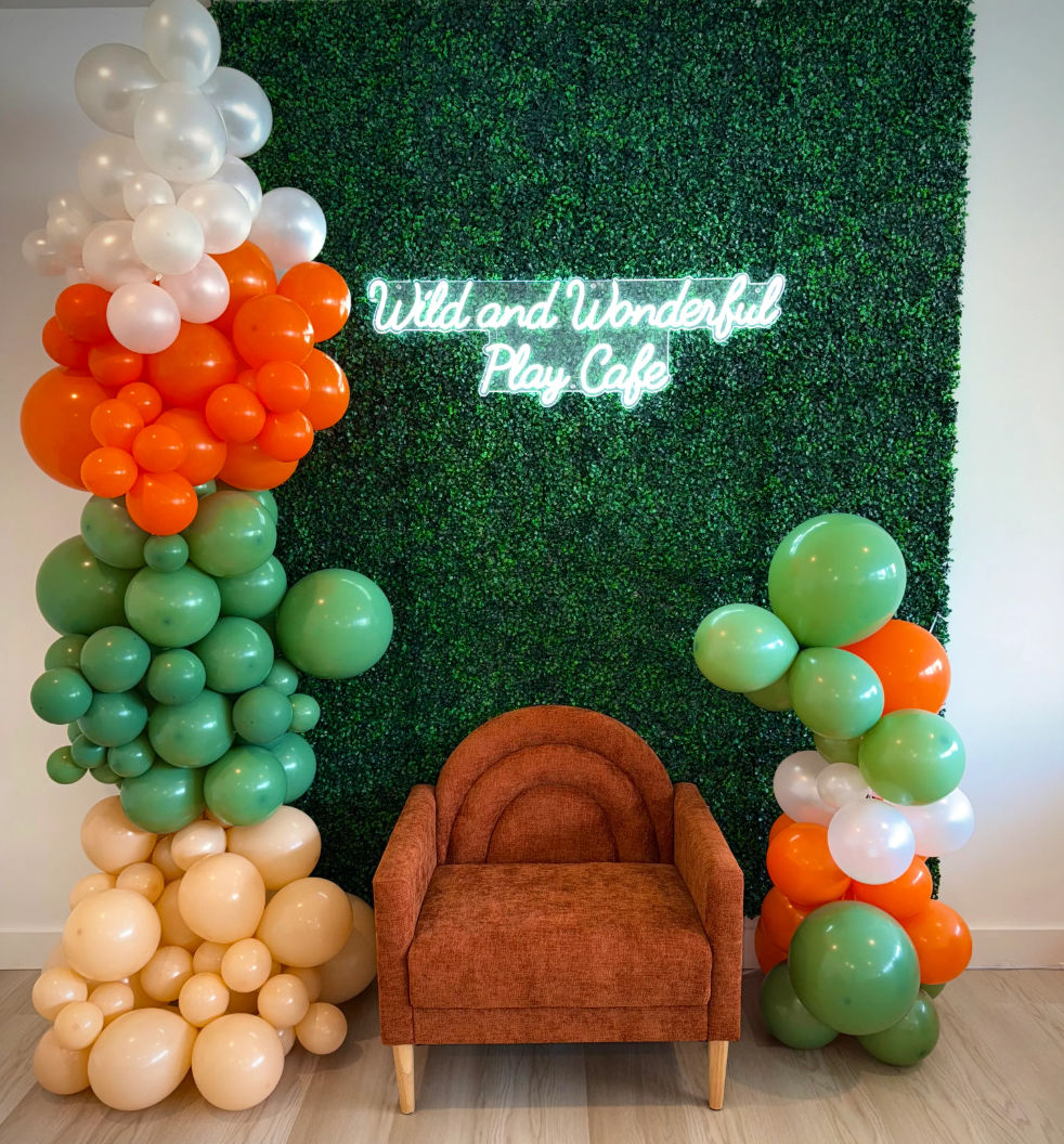 Playful photo backdrop: rust-orange armchair in front of a green boxwood wall with a neon script sign, flanked by green, orange, cream and white balloon garlands — kids play-cafe seating area.