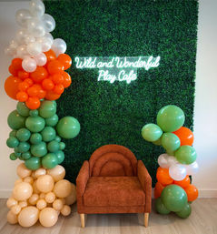 Playful photo backdrop: rust-orange armchair in front of a green boxwood wall with a neon script sign, flanked by green, orange, cream and white balloon garlands — kids play-cafe seating area.