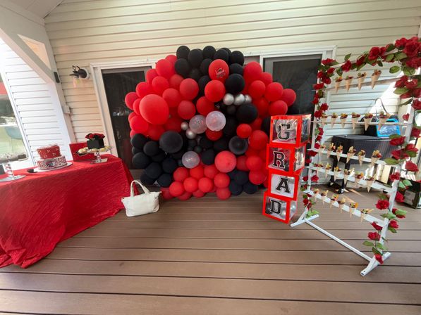 Red and black balloon wall and stacked GRAD blocks on a covered backyard patio, red-draped dessert table with graduation cake and a rose-adorned rack of hanging snack cones.