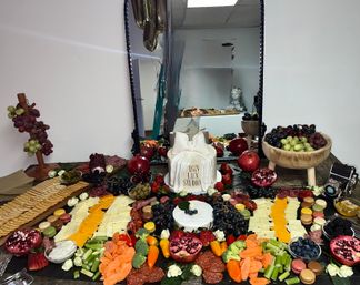 Charcuterie grazing table in a studio with assorted cheeses, cured meats, crackers, grapes, pomegranate halves, berries, veggies, macarons and a decorative white bow-topped cake reflected in a mirror.