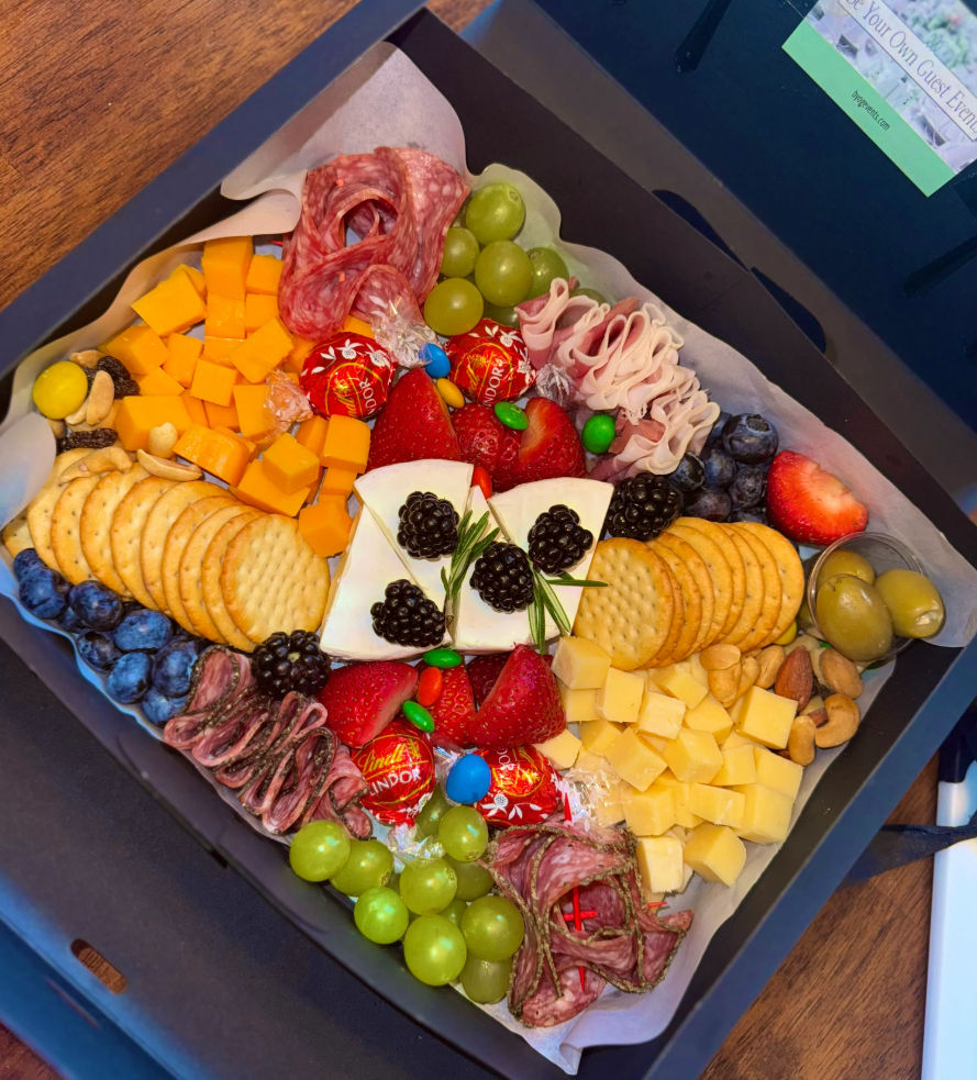 Colorful charcuterie gift box: crackers, cubed cheddar and Swiss, Brie topped with blackberries and rosemary, sliced salami and ham, green grapes, strawberries, blueberries, olives, mixed nuts, and wrapped chocolate truffles.