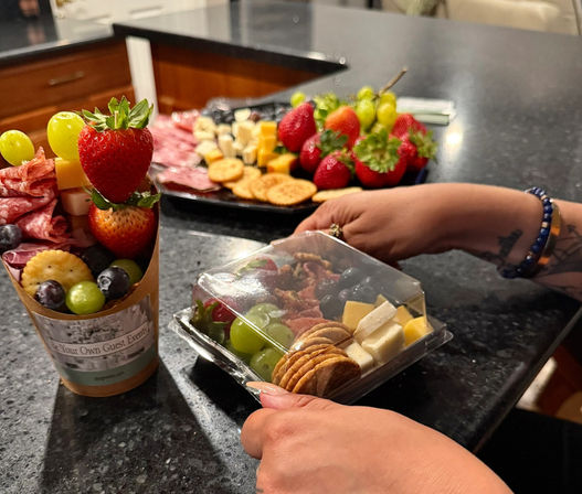 Hands placing a grab-and-go charcuterie box on a dark kitchen countertop beside a paper charcuterie cup and platter of strawberries, grapes, crackers, cheeses and cured meats.