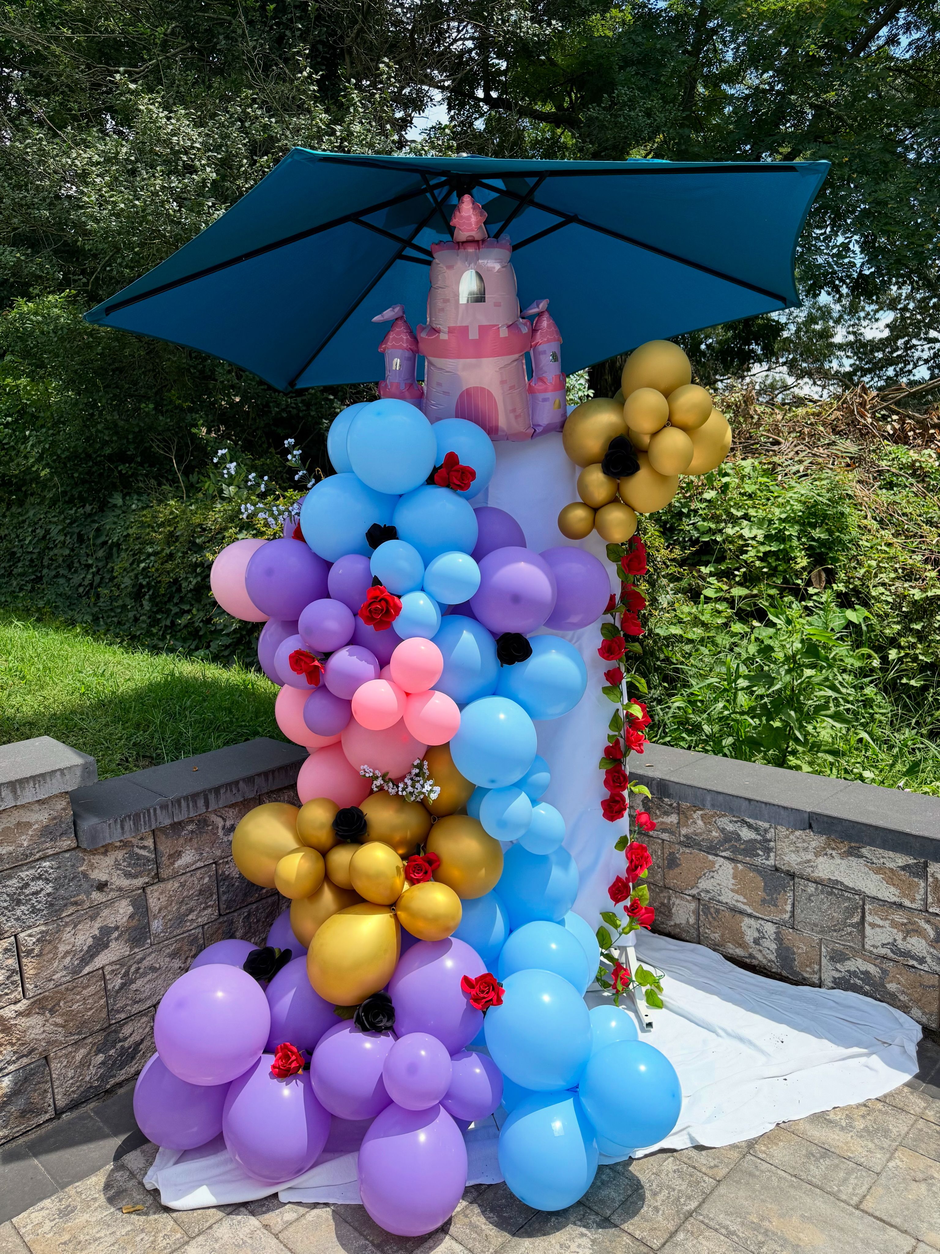 Whimsical princess balloon column with inflatable pink castle topper under a blue umbrella — pastel blue, purple, pink and gold balloons with red rose accents on a stone patio in a backyard garden for an outdoor birthday party