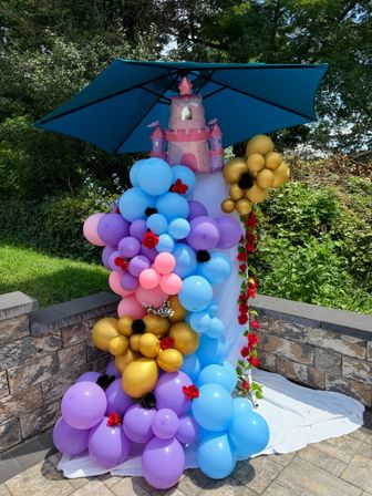 Whimsical princess balloon column with inflatable pink castle topper under a blue umbrella — pastel blue, purple, pink and gold balloons with red rose accents on a stone patio in a backyard garden for an outdoor birthday party