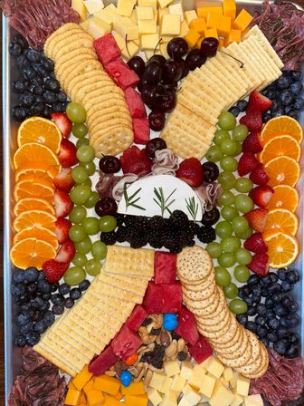 Vibrant charcuterie tray with crackers, cubed cheeses and a rosemary-topped wheel of brie with blackberries, surrounded by orange slices, green grapes, strawberries, blueberries, cherries, watermelon, salami, mixed nuts and candies — festive appetizer board.