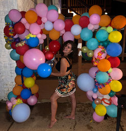 Smiling person posing barefoot under a vibrant balloon arch at a nighttime backyard party, wearing a tropical-print skirt and black top, surrounded by multicolored balloons and floral leis.