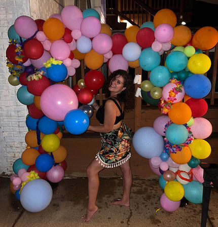 Smiling person posing barefoot under a vibrant balloon arch at a nighttime backyard party, wearing a tropical-print skirt and black top, surrounded by multicolored balloons and floral leis.