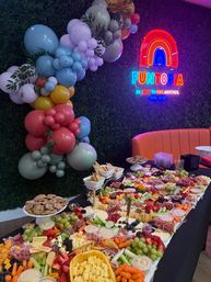 Colorful indoor party setup with a neon rainbow sign, pastel balloon garland and a large grazing charcuterie table of cheeses, fruits, crackers and dips.