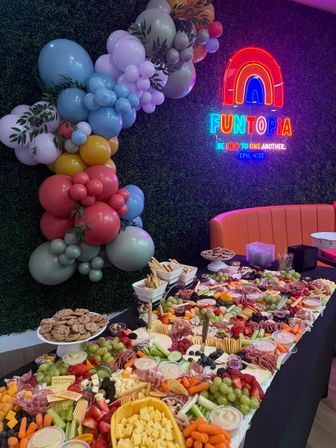Colorful indoor party setup with a neon rainbow sign, pastel balloon garland and a large grazing charcuterie table of cheeses, fruits, crackers and dips.
