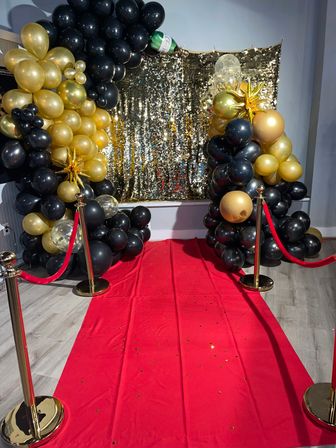 Indoor red carpet photo booth with shimmering gold sequin backdrop, black-and-gold balloon arch, gold stanchions with red ropes and scattered confetti