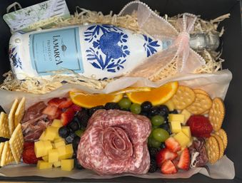 Decorative gift box with a bow-tied sparkling wine bottle on shredded paper above a charcuterie spread—salami shaped into a rose, cubed cheese, strawberries, grapes, blackberries, orange slices and round crackers.