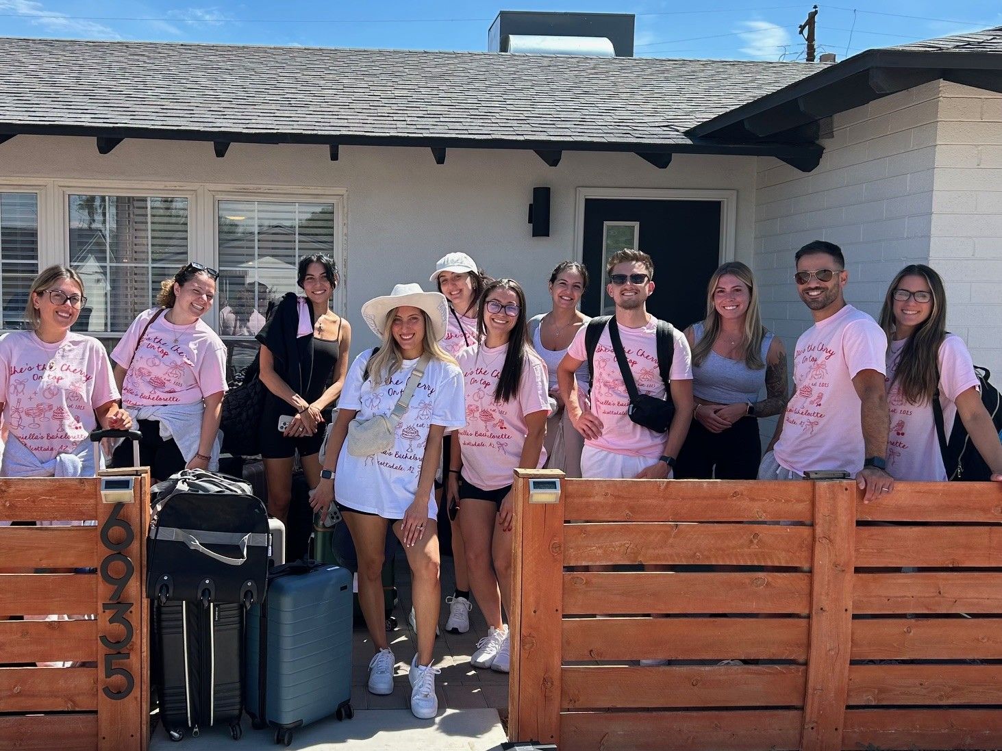 Smiling group of friends in matching pink bachelorette T‑shirts and summer outfits standing outside a single‑story rental house with luggage by a wooden gate on a sunny day