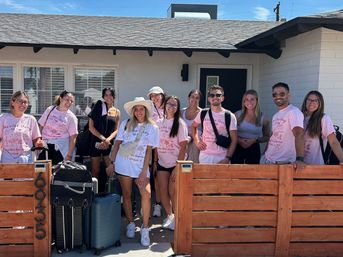 Smiling group of friends in matching pink bachelorette T‑shirts and summer outfits standing outside a single‑story rental house with luggage by a wooden gate on a sunny day