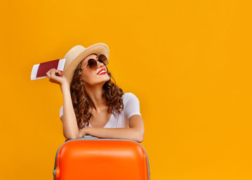 Smiling woman in a straw hat and sunglasses leaning on a bright orange suitcase, holding a passport and boarding pass against a vibrant yellow background — summer vacation travel concept.