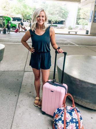 Smiling woman in a black romper at an airport curbside holding a pink rolling carry-on and a navy floral duffel, outdoor terminal pickup area ready to travel