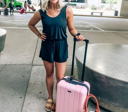 Smiling woman in a black romper at an airport curbside holding a pink rolling carry-on and a navy floral duffel, outdoor terminal pickup area ready to travel