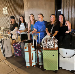 Six smiling women with colorful suitcases and duffel bags waiting at an airport curbside pickup at night, posed in front of a textured concrete wall.