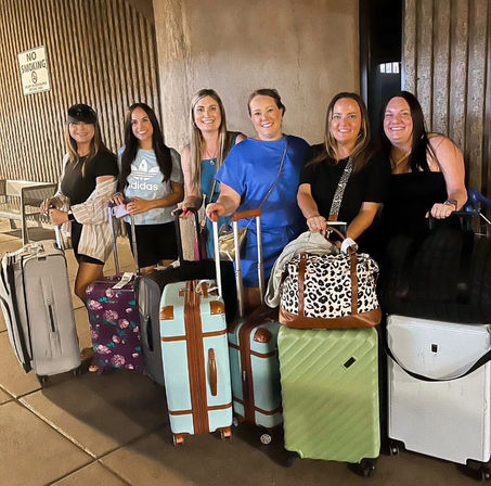 Six smiling women with colorful suitcases and duffel bags waiting at an airport curbside pickup at night, posed in front of a textured concrete wall.