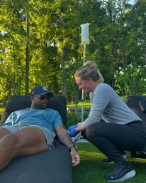 Outdoor IV therapy scene: man in sunglasses relaxed on a lounge chair in a sunlit backyard while a smiling clinician kneels inserting an IV into his tattooed arm with the fluid bag on a pole, surrounded by green trees and white flowers.