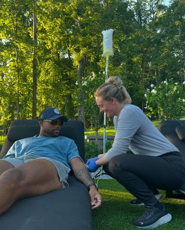 Outdoor IV therapy scene: man in sunglasses relaxed on a lounge chair in a sunlit backyard while a smiling clinician kneels inserting an IV into his tattooed arm with the fluid bag on a pole, surrounded by green trees and white flowers.