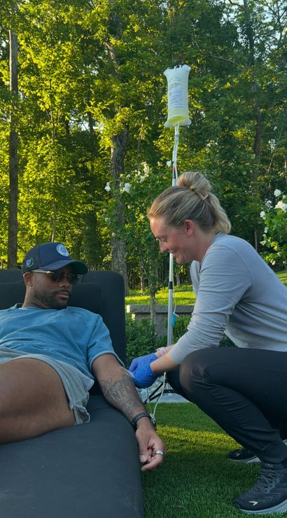 Outdoor IV therapy scene: man in sunglasses relaxed on a lounge chair in a sunlit backyard while a smiling clinician kneels inserting an IV into his tattooed arm with the fluid bag on a pole, surrounded by green trees and white flowers.