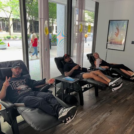 Three adults reclining in black lounge chairs receiving IV therapy from yellow IV bags inside a bright downtown IV therapy lounge with large street-facing windows; two people smile and one flashes a peace sign.