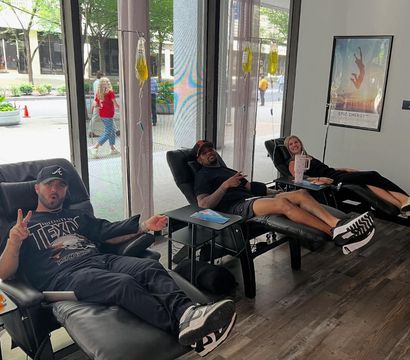 Three adults reclining in black lounge chairs receiving IV therapy from yellow IV bags inside a bright downtown IV therapy lounge with large street-facing windows; two people smile and one flashes a peace sign.