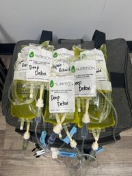 Portable IV therapy kit with multiple yellow-green nutrient bags labeled "Deep Detox" and connected tubing on a gray carrying case in a treatment room.