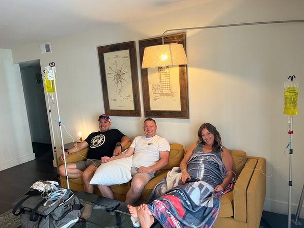 Three smiling adults on a yellow sofa in a bright living room receiving IV hydration drips, one wrapped in a blanket; IV poles, tubing and travel bags visible on a glass coffee table.