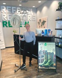 Smiling woman in a modern IV therapy clinic reception holding an IV pole topped with a cowboy hat, with bold “Refresh Restore Renew” wall signage and a poster featuring green cowboy boots.