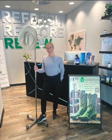 Smiling woman in a modern IV therapy clinic reception holding an IV pole topped with a cowboy hat, with bold “Refresh Restore Renew” wall signage and a poster featuring green cowboy boots.