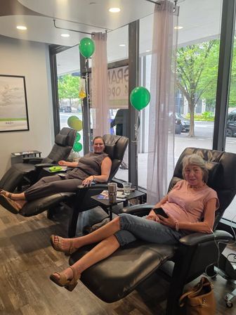 Two people reclining in black leather chairs at a wellness infusion lounge with IV bags, green balloons, and floor-to-ceiling windows overlooking a tree-lined city street.
