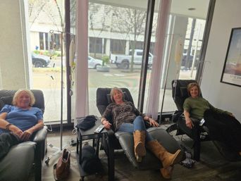 Three women reclining in black lounge chairs receiving IV therapy in a bright wellness infusion lounge with floor-to-ceiling windows and an urban street view
