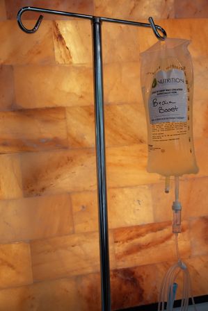 Stainless steel IV stand with a 'Brain Boost' nutrition IV drip bag and tubing hanging against a glowing Himalayan salt wall — wellness IV therapy infusion setup at a spa/clinic.