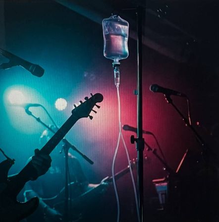 Silhouette of electric guitar and microphones on a live music stage with an IV drip prop, bathed in teal and magenta concert lights