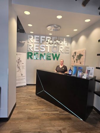 Bright modern wellness clinic reception lobby with black angular front desk, staff behind counter, hanging pendant lights and large wall lettering reading 'REFRESH RESTORE RENEW'.