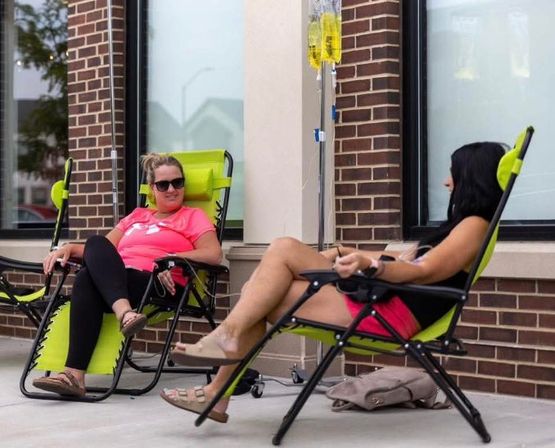 Two people lounging in lime-green recliners on a brick storefront patio, chatting while receiving IV infusions from yellow bags