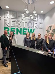 Smiling reception team posing behind a black front desk in a modern wellness reception area with large green and white wall letters reading REFRESH RESTORE RENEW, hanging pendant lights and a wall map.