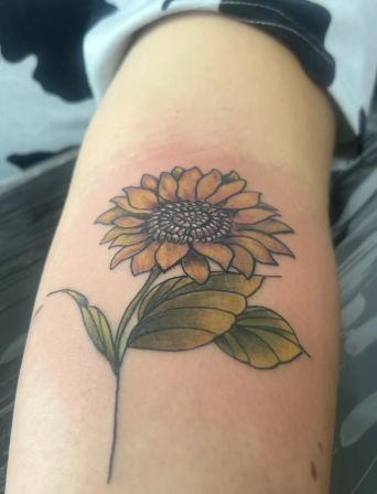 Close-up of a fresh sunflower tattoo on an inner forearm with yellow-orange petals, detailed textured center, green shaded leaves and a thin black stem