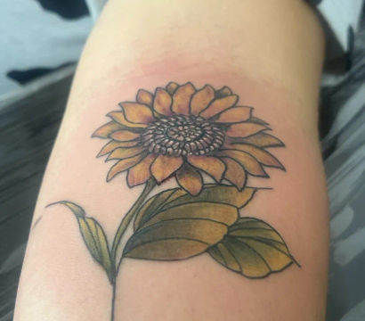 Close-up of a fresh sunflower tattoo on an inner forearm with yellow-orange petals, detailed textured center, green shaded leaves and a thin black stem
