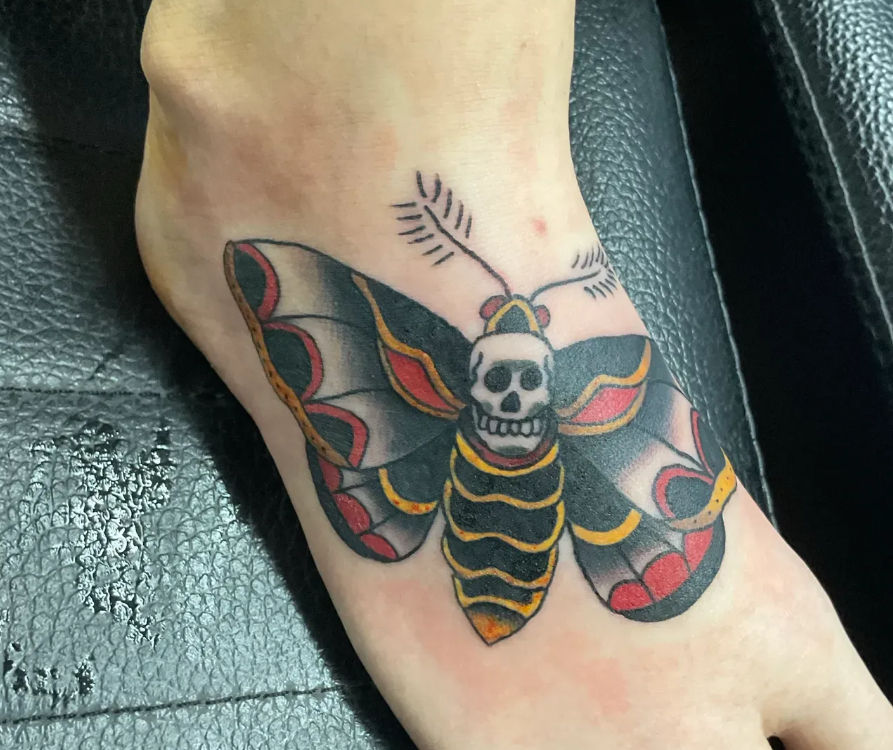 Bold traditional-style foot tattoo of a skull-faced moth across the ankle, featuring black wings with red and yellow accents.