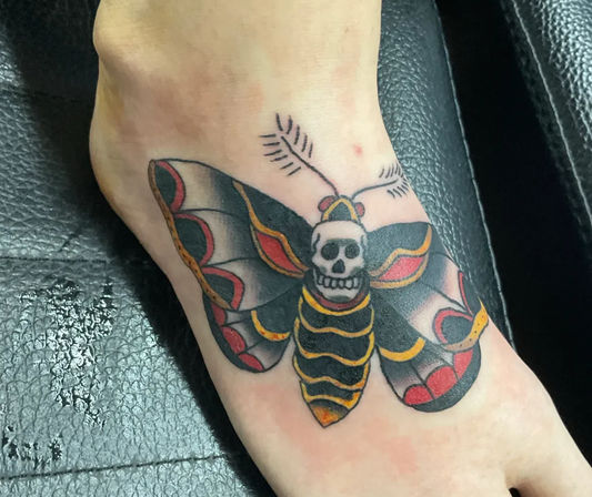 Bold traditional-style foot tattoo of a skull-faced moth across the ankle, featuring black wings with red and yellow accents.