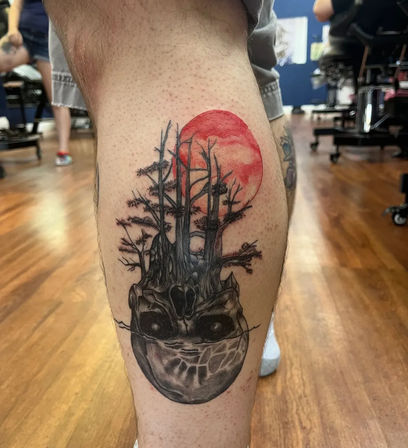 Calf tattoo of a surreal skull that morphs into leafless trees beneath a red full moon — black-and-gray shading with a red accent on a lower leg in a tattoo studio setting.