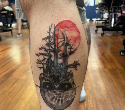 Calf tattoo of a surreal skull that morphs into leafless trees beneath a red full moon — black-and-gray shading with a red accent on a lower leg in a tattoo studio setting.