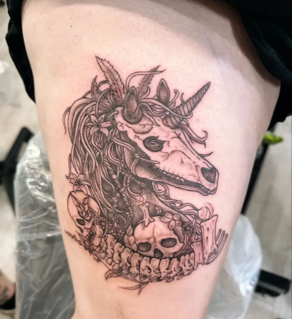 Black-and-grey thigh tattoo of a macabre unicorn skull with flowing mane, flowers and small human skulls woven into a spinal vertebra border, detailed linework fantasy-horror design.